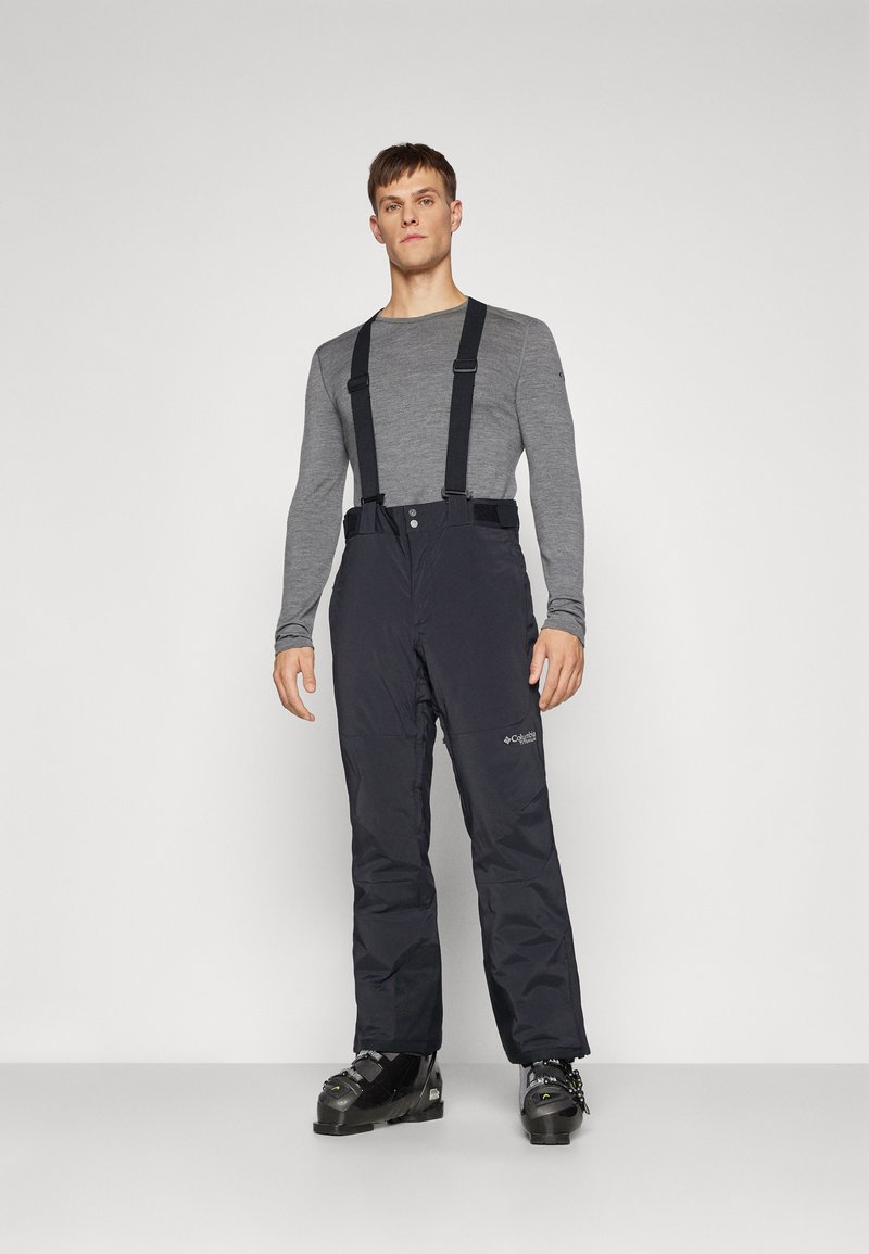 Pantalon de ski noir avec bretelles réglables, fabriqué en matériau imperméable. Présente un détail de logo sur la jambe. Porté avec des bottes de ski.