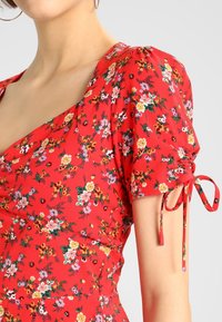 Rote Bluse mit floralem Muster, kurzen Puffärmeln, elastischen Akzenten und einer Bindeschnur. Verziert mit bunten Blumenmustern auf einem strukturierten Stoff.