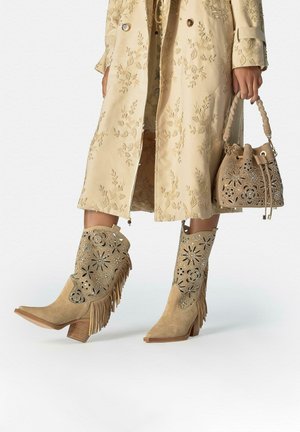 Gabardina beige bordada con botas de ante con flecos y recortes florales a juego, y un pequeño bolso tipo cubo con asa trenzada.