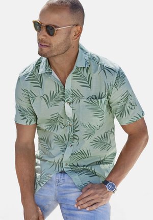Helles grünes tropisches Hemd mit Blattmuster, kurzen Ärmeln und einer Brusttasche; kombiniert mit blauen Jeans und Sonnenbrille.