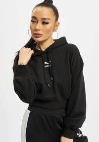 Sudadera negra recortada hecha de un tejido suave, con capucha ajustable y un logo blanco de Puma en el pecho, combinada con pantalones de chándal negros.