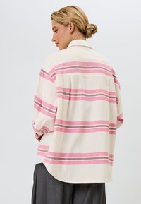 Oversized button-up skjorte i lys creme med rosa og grå vandrette striber. Har en rygkappe og en buet søm.