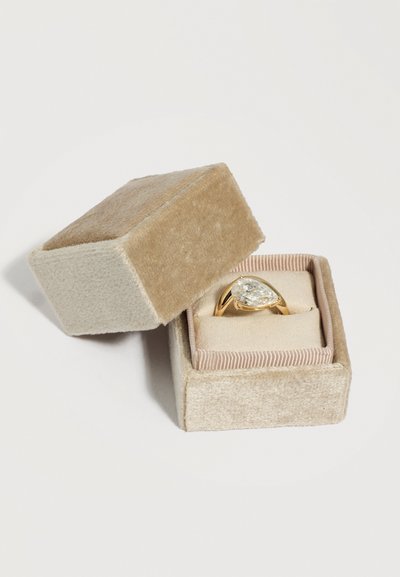 Ανοιχτό μπεζ βελούδινο κουτί δαχτυλιδιού που αποκαλύπτει χρυσό δαχτυλίδι με μεγάλο αχλαδόσχημο διαυγές πολύτιμο λίθο πάνω σε μαλακό μαξιλάρι.