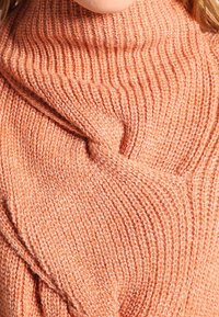 Detailní záběr na osobu v texturovaném, žebrovaném pleteném svetru v jemně broskvové barvě s vysokým límcem.