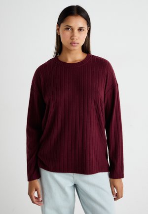 Bordeauxroter gerippter Pullover mit rundem Ausschnitt und langen Ärmeln, in lockerer Passform und weichem Material, kombiniert mit hellblauen Jeans.