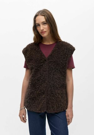 Femme aux longs cheveux bruns portant une veste duveteuse marron foncé sur un t-shirt bordeaux à manches courtes et un jean bleu, debout devant un fond blanc.