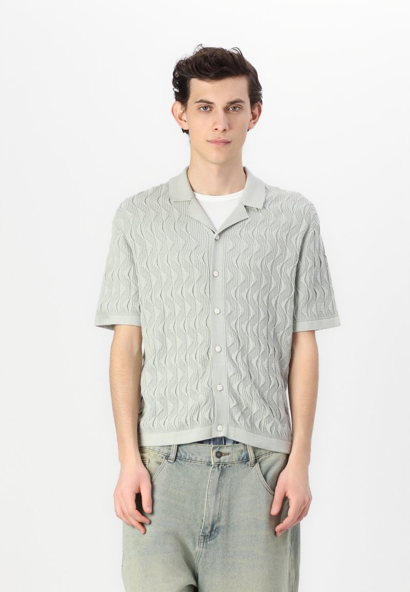 Camisa de manga corta de color verde claro con un patrón de olas, botones en la parte frontal y cuello plano. Hecha de tela texturizada, combinada con jeans claros.