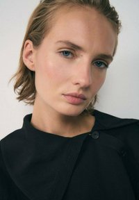 Schwarze Jacke mit großem Kragen und Knopfverschluss, getragen von einem Modell mit kurzen blonden Haaren, heller Haut und blauen Augen.