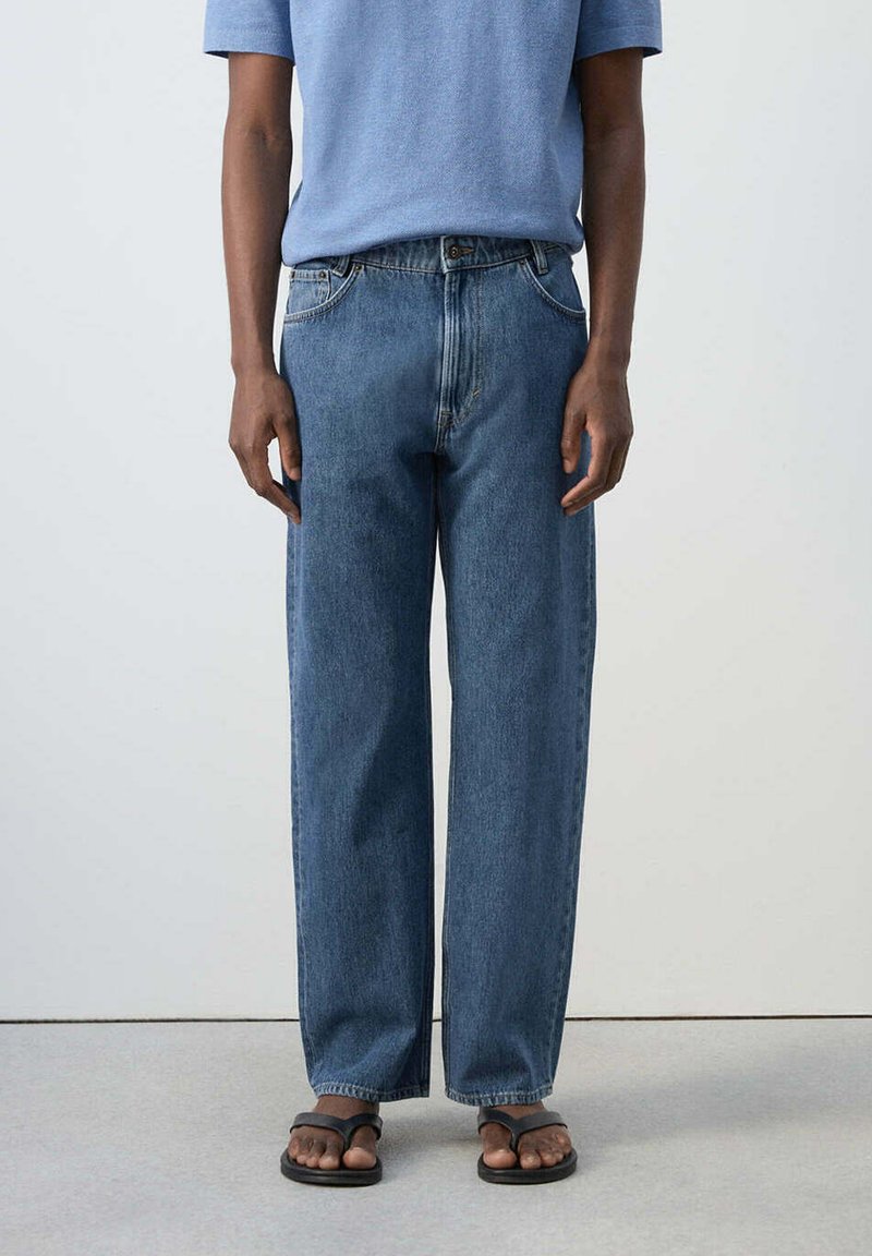 Personne portant un jean bleu ample, une chemise bleu clair à manches courtes et des tongs noires, debout devant un fond blanc uni.