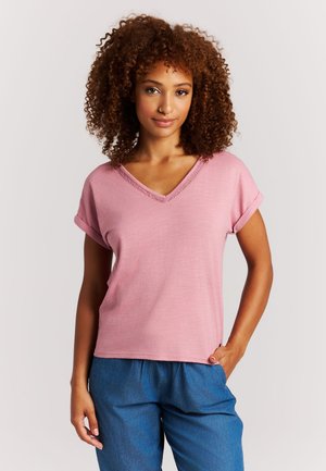 Vrouw met krullend haar, gekleed in een roze V-hals top met korte mouwen en een blauwe broek, staand met één hand in haar broekzak tegen een effen achtergrond.