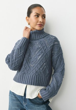Pulover tricotat de culoare albastră închisă, scurtat, cu guler înalt, având un model în diamante și manșete cu rădăcină, purtat peste o cămașă albă cu guler.