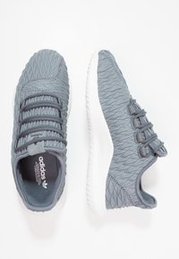 Grå sportskor med en texturerad ovandel, med ett mönstrat design, elastiska snören och en vit gummisula. Adidas-logotyp synlig.