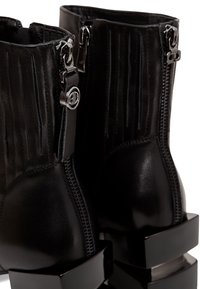 Stivaletti alla caviglia in pelle nera con pieghe verticali, cerniere argentate, dettagli con logo e tacchi spessi neri lucidi. Texture liscia, design moderno.