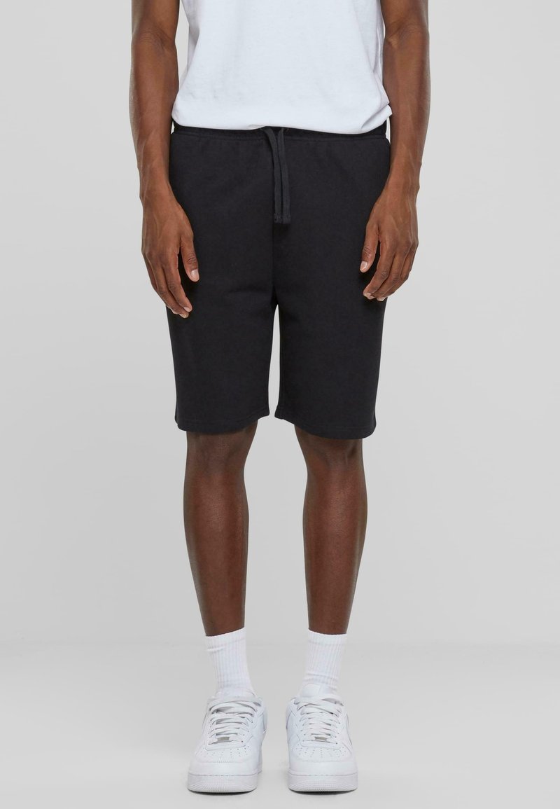 Homme portant un short noir à cordon, des chaussettes blanches, des baskets blanches et une chemise blanche, debout devant un fond gris.