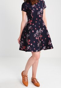 Žena v námořnickém květinovém šatech s krátkými rukávy a hnědými šněrovacími balerínami, stojící se zkříženýma nohama.