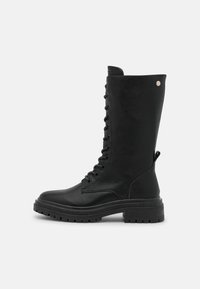 Černá kožená bojová bu boot do výšky kolen vyrobená z hladké kůže, s předním šněrováním, robustní podrážkou a kovovým akcentem blízko vršku.