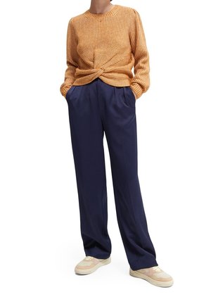 Oranje gebreide trui met een gedraaide voorkant, gecombineerd met marineblauwe wijde broekspijpen en beige sneakers met roze accenten.