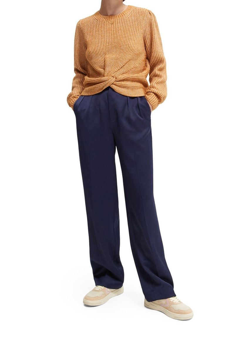 Pull en maille orange avec un design torsadé à l'avant, associé à un pantalon large bleu marine et des baskets beiges avec des touches de rose.