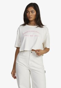 White cropped t-shirt with pink printed text, "SUNSETS AND MISERY CALL 1800 - RVCA." Short sleeves, soft texture, relaxed fit.