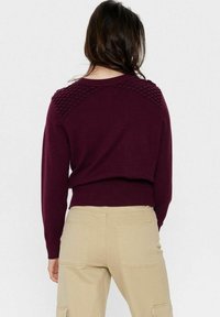 Bordowy sweter z teksturowanymi akcentami na ramionach i prążkowanym dołem, wykonany z dzianiny. Noszony z beżowymi spodniami, widok z tyłu.