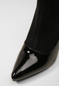 Chaussure à talon haut noire à bout pointu avec une finition en cuir verni brillant et une tige en tissu cousu.