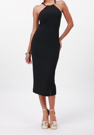 Frau trägt ein ärmelloses schwarzes Midikleid mit Neckholder und goldene Plateau-Pumps, steht mit einer Hand in der Hüfte.