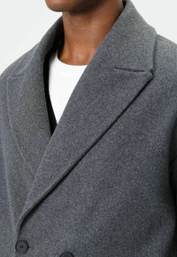 Dubbele rij grijze wollen jas met een kraag met inkeping, gestructureerde stof en zwarte knopen, gedragen over een wit overhemd.