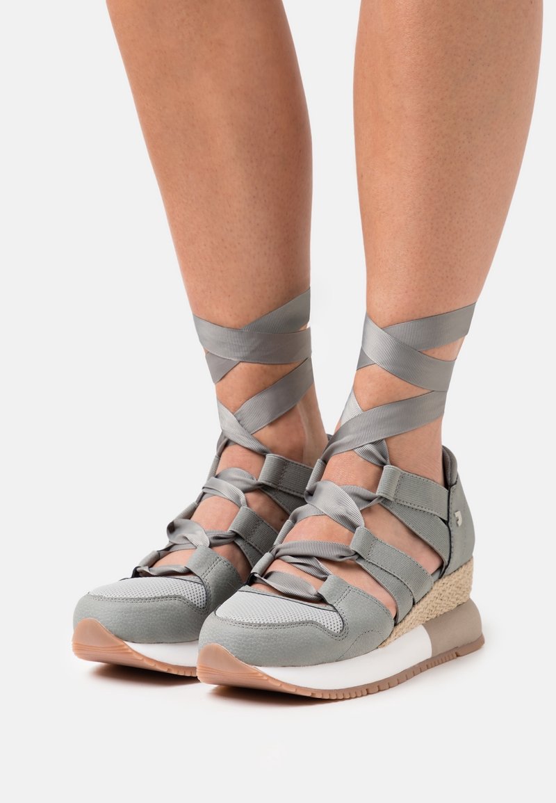 Pieds portant des baskets compensées grises avec des rubans croisés aux chevilles, noués au-dessus des chevilles sur fond blanc.