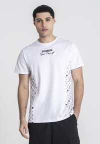 Bílá bavlněná tričko s černým logem na hrudi a vzorovaným bočním designem s ikonami a iniciály na obou stranách.