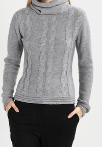 Jersey gris de punto con cuello de tortuga, que presenta un patrón de trenzas texturizado, mangas largas y un diseño ajustado. La tela tiene una textura suave.