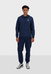 Completo felpa e pantaloni jogging blu navy in morbido tessuto, con tasca a canguro, cappuccio con cordino e dettaglio del logo sul petto.