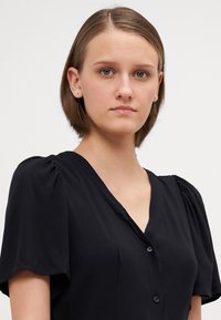 Blusa nera a maniche corte con scollatura a V, chiusura a bottoni e maniche arricciate, realizzata in un tessuto liscio con una leggera lucentezza.