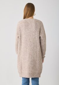 Pull oversized beige clair avec une texture douce et duveteuse, des détails côtelés au niveau du col et des poignets, et des fentes sur les côtés pour faciliter le mouvement.