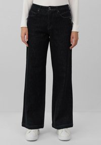 Pantalons en denim noir à jambes larges avec une texture subtile, design à cinq poches, fermeture à bouton en métal et couture contrastante. Associés à des baskets blanches.