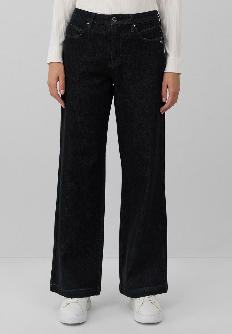 Pantalons en denim noir à jambes larges avec une texture subtile, design à cinq poches, fermeture à bouton en métal et couture contrastante. Associés à des baskets blanches.