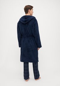 Marineblå hettebadekåpe med teksturert, myk stoff. Har belte med knyting og sidelommer, kombinert med rutete pysjamasbukser.