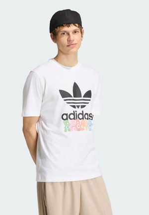 Jeune homme portant un t-shirt blanc Adidas avec logo noir et oursons colorés, pantalon beige et casquette noire à l'envers.