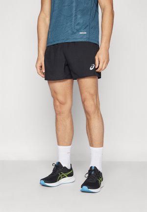 Mężczyzna noszący czarne spodenki do biegania Asics, białe skarpetki, czarno-zielone buty do biegania oraz niebieską sportową koszulkę stoi na jednolitym tle.