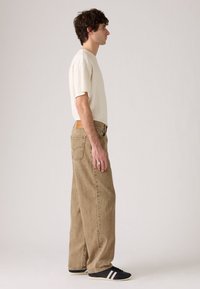 Beige t-shirt met korte mouwen, gecombineerd met losse, lichtbruine jeans. Het onderwerp draagt zwarte sneakers met witte strepen en een riem van gestructureerde stof.