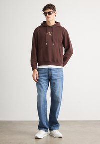 Brun hettegenser med frontlomme og logo, kombinert med lyseblå jeans og hvite sneakers. Modell iført mørke solbriller.