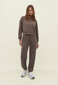 Bruine, lange mouw sweatshirt met een cropped fit en een trekkoord in de taille, gecombineerd met bijpassende tapered sweatpants. G gedragen met zilveren sportschoenen.