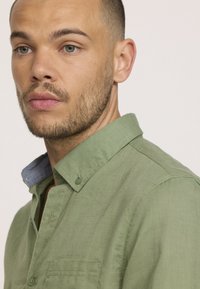 Chemise boutonnée en lin vert avec un col, équipée d'une bande intérieure de col ressemblant à du denim ; comprend une poche à l'avant et des détails de couture visibles.