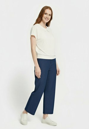 Lächelnde Frau mit roten Haaren, die ein weißes Kurzarmoberteil, marineblaue weit geschnittene Hose und weiße Turnschuhe trägt, vor einem schlichten Hintergrund stehend.