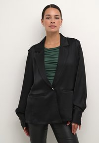 Blazer negru din satin cu o croială lejeră, închidere cu un singur nasture și două buzunare în față. Purtați deasupra unei bluze verzi texturate. Textură netedă și elegantă.
