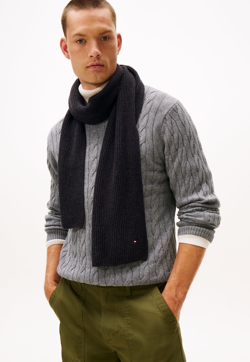 Mann trägt einen grauen Kabelstrickpullover, einen dunklen Rippschal und olivgrüne Hose mit Händen in den Taschen vor weißem Hintergrund.