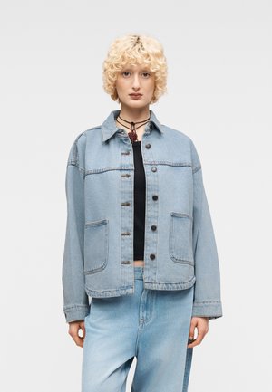 Göndör szőke hajú személy világoskék, túlméretezett farmerruhával, fekete felsővel és világoskék farmernadrággal, egyszínű háttér előtt állva.