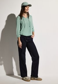 Blouse vert clair avec un décolleté en V et des cordons, jeans noirs et baskets beiges, complétée par une casquette assortie.