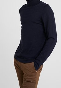 Pull à col roulé bleu marine en tissu doux, avec manches longues et coupe ajustée. Associé à un pantalon de couleur beige.