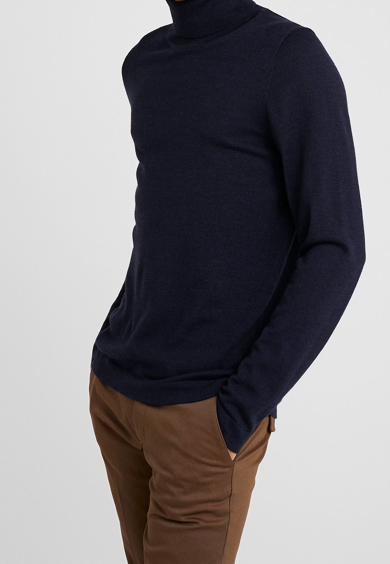 Pull à col roulé bleu marine en tissu doux, avec manches longues et coupe ajustée. Associé à un pantalon de couleur beige.