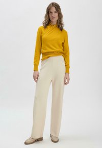Geel wikkelsweater met lange mouwen, gecombineerd met crèmekleurige wijde broek en beige schoenen. Zachte textuur en minimalistisch ontwerp.